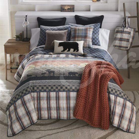 A front view of the quilt on a bed. The quilt has a sun on a mountain horizon with wildlife silhouettes on a pine backdrop, along with plaid and gingham patterns.